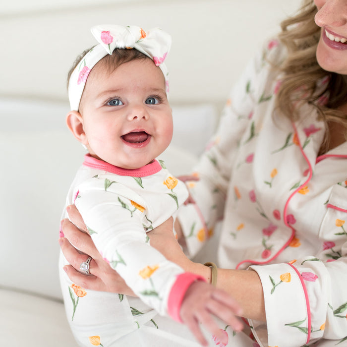 Close up of infant wearing the Zippered Romper in Tulip with matching knotted bow headband being held by her mother matching in pajamas sitting on a bed